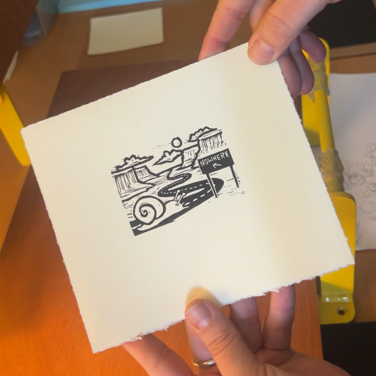 Handmade mini linocut print of a snail on a winding desert road to nowhere in black ink on warm white paper