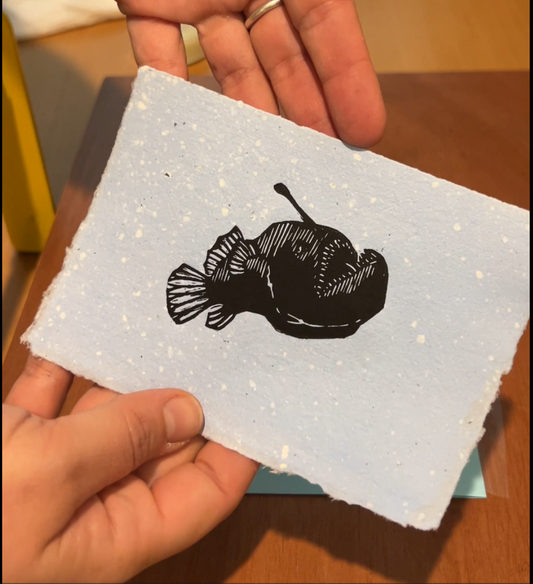 Handmade mini linocut print of an angler fish in black ink on blue speckled handmade paper